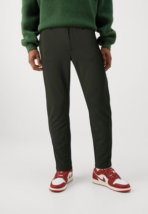 Pantalons vert foncé à coupe ajustée, texture lisse, avec plis sur le devant, associés à des baskets rouges et blanches au design classique.