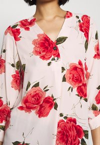 Blouse fleurie avec un fond rose pâle, de grandes roses rouges, des feuilles vertes, un décolleté en V et trois boutons sur le devant.