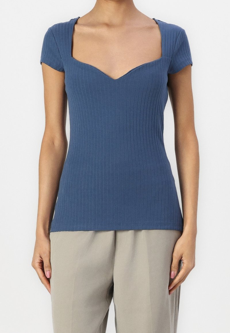 Femme portant un haut bleu côtelé à manches courtes avec un décolleté en cœur et un pantalon beige clair, debout devant un fond uni.
