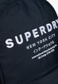 Czarny nylonowy kapelusz z białym haftowanym napisem "SUPERDRY" i "NEW YORK CITY", z dodatkowym tekstem w języku japońskim oraz szczegółami produkcji.