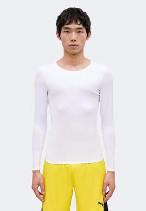 Ung mand, der står med fronten mod kameraet, iført en tætsiddende hvid langærmet skjorte og lyse gule sportsshorts.