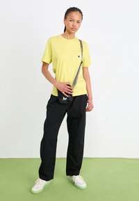 Gelbes T-Shirt mit kurzen Ärmeln und Brusttasche, kombiniert mit schwarzen lockeren Hosen und einer kleinen schwarzen Umhängetasche. Weiße Sneakers mit grünen Sohlen.