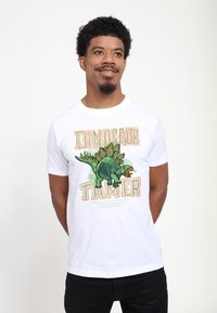 Muž stojící s rukama za zády, oblečený v bílém tričku se zeleným dinosaurem a textem "Dino tamer" na přední straně.