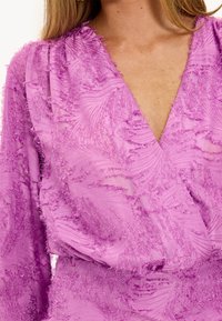 Blouse lavande avec un décolleté en V profond, présentant un tissu texturé avec des détails effilochés et des manches longues, mettant en valeur un design fluide.