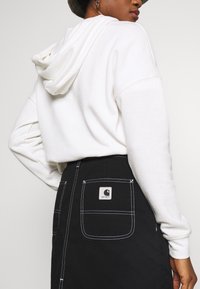 White hoodie with a drawstring hood, paired with a black skirt featuring white stitching and a square logo patch on the back pocket.