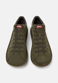 Olivově zelené tenisky s texturovanou semišovou úpravou, s červeným logem a elastickými tkaničkami, umístěné na neutrálním pozadí.