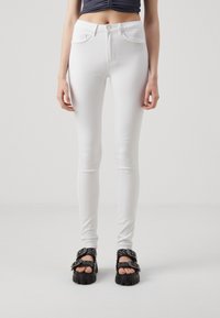 Jeans blancos ajustados con diseño de cinco bolsillos, cierre de botón y textura suave, combinados con sandalias negras gruesas con detalles de tachuelas.