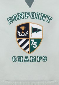 Felpa grigia con un design ricamato a scudo che presenta un trofeo, una bandiera e una corona di alloro, accentuato da elementi verdi e dorati. Il testo recita "BONPOINT CHAMPS."