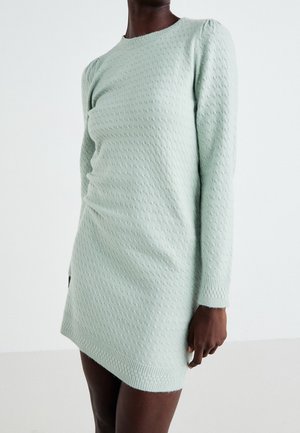 Femme portant une robe pull en maille texturée vert clair avec des manches longues et un ourlet asymétrique, debout sur un fond uni.