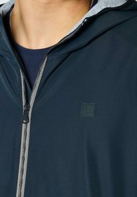 Námořnická kapucová bunda se zipem, s šedou vnitřní podšívkou, kontrastním šitím a texturovanou nášivkou s logem na hrudi.