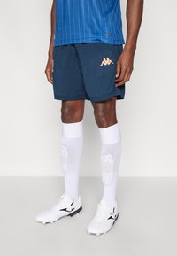 Peach színű tengerészkék sportnadr�ág Kappa logóval, fehér térdig érő zoknival és fehér, fekete részletekkel díszített futballcipővel párosítva.