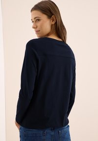 Navy blue long-sleeve top with a round neckline and a seam detail across the back, paired with denim jeans. The fabric appears soft and smooth.