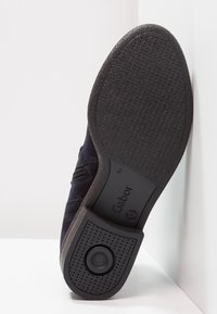 Svart mocka ankelsko med texturerad gummisula, sidodragkedja och präglad varumärkeslogotyp. Har en cirkulär detalj på hälen.