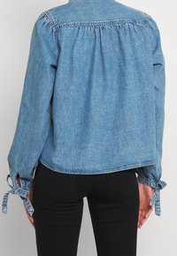 Vista trasera de una persona con una camisa de mezclilla de manga larga azul claro, con yugo fruncido y puños atados, combinada con pantalones negros.