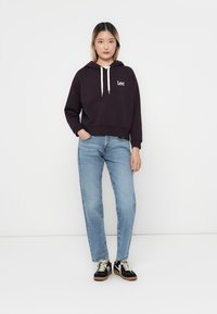 Haut à capuche violet foncé avec cordons et logo blanc, associé à un jean bleu clair et des baskets noires avec des accents blancs. Arrière-plan uni.