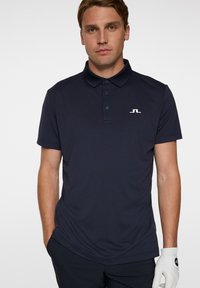 Marinblå pikétröja i andningsbart material, med klassisk krage, tre-knapps knäppning och en liten vit logotyp på bröstet.