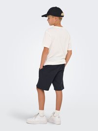 Hvid T-shirt, sorte shorts og hvide sneakers med strukturerede såler; en sort kasket fuldender det afslappede outfit. Fokus på den løse pasform og farver.
