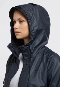 Chaqueta de lluvia marina con capucha, hecha de material suave y resistente al agua. Cuenta con una cremallera, botones y un logo en el pecho.