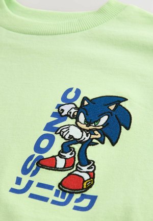 Vaaleanvihreä t-paita, jossa on brodeerattu sininen hahmo määrätietoisella ilmeellä, punaiset kengät ja sinisellä tyylitelty teksti "SONIC".