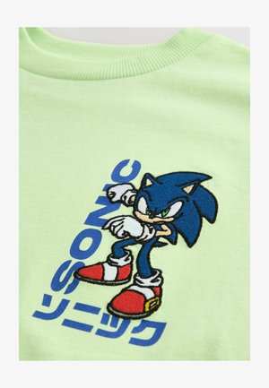 Jasnozielona koszulka z haftowanym niebieskim bohaterem o zdeterminowanym wyrazie twarzy, czerwonymi butami i niebieskim napisem "SONIC" w stylizowanej czcionce.
