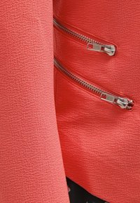 Veste texturée corail avec trois fermetures éclair argentées sur le devant, dotée d'un tissu intérieur lisse contrastant avec la surface extérieure.