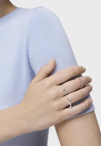 Anillos de plata con varios diseños en una mano apoyada sobre una tela de punto suave de color azul claro. La tela tiene una textura lisa y una forma ajustada.