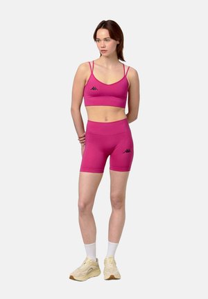 Femme debout, mains dans le dos, portant un soutien-gorge de sport rose et un short assorti, des chaussettes blanches et des baskets beiges sur fond clair.