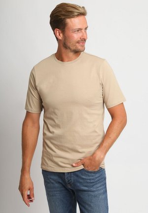 Blonder Mann, der ein beiges Kurzarmhemd und blaue Jeans trägt, steht mit einer Hand in der Hosentasche und schaut nach rechts vor schlichtem Hintergrund.