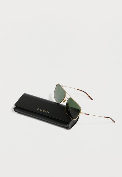 Χρυσά γυαλιά ηλίου aviator με πράσινους φακούς, τοποθετημένα πάνω σε μαύρη θήκη Gucci με χρυσό λογότυπο, σε λευκό φόντο.