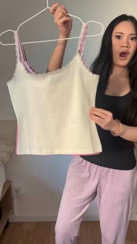 Vrouw met lang donker haar houdt een crèmekleurig hemdje met roze kanten bandjes aan een hanger vast, draagt een zwart topje en roze broek, en toont verrassing.
