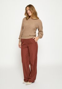 Gebreide beige trui met een gestructureerd patroon, gecombineerd met roestkleurige, vloeiende broek. Model staat met handen in de zakken en draagt witte, puntige schoenen.