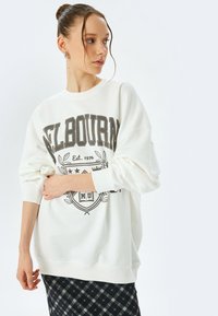 Sudadera blanca con gráfico "MELBOURNE" en marrón oscuro, con escote redondeado, mangas largas y puños de canalé. Combínala con una falda de cuadros.