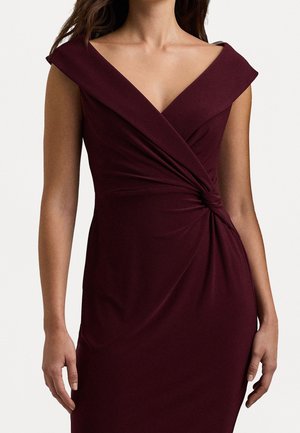 Kvinde iført ærmeløs, off-shoulder bordeaux kjole med knudedetalje ved taljen, foran en ensfarvet lys baggrund.