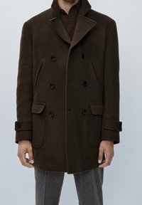 Cappotto doppio petto marrone in lana con grandi bottoni, tasche a patta e colletto alto, indossato sopra un maglione marrone a coste e pantaloni grigi.