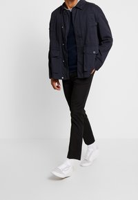 Man som bär en marinblå jacka, svarta byxor, vita sneakers och vita strumpor som går mot en helt vit bakgrund.