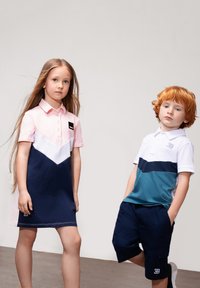 Due bambini indossano abiti contrastanti: un vestito polo rosa e blu navy con motivo chevron bianco e una polo a maniche corte con blocchi di colore orizzontali in bianco, teal e blu navy.