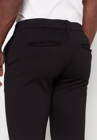 Zwarte broek met een slim fit, twee achterzakken, een gladde textuur en subtiele stiksels langs de taille.