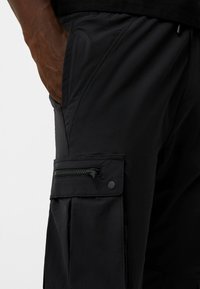 Svarta cargoshorts gjorda av lättviktsmaterial, med en sidoficka med dragkedja och tryckknappsstängning, samt sömnadsdetaljer på sömmarna.