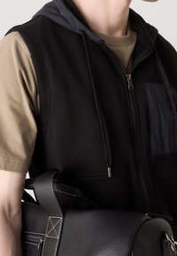 Osoba v černé zipové vestě přes hnědou košilku s krátkým rukávem, nese černou koženou tašku s bílým prošíváním na popruhu.