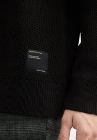 Schwarzer, gestrickter Pullover mit einer strukturierten Oberfläche, ausgestattet mit einem gerippten Saum und einem eingewebten Markenetikett an der Seite. Sichtbar sind kontrastierende Karohosen.