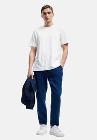 Weißes Baumwoll-T-Shirt, strukturierten marineblauen Hosen, hält eine marineblaue Jacke und trägt weiße Sneakers, steht vor einem neutralen Hintergrund.