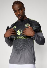 Schwarz langärmliges Trikot mit gradientem Graumuster, grünen Akzenten und dem Logo von Manchester United. Hält schwarze Fußballschuhe in beiden Händen.