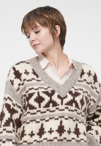 Jeune personne avec les cheveux bruns courts portant une chemise blanche à col sous un épais pull tricoté à motifs beige et marron.