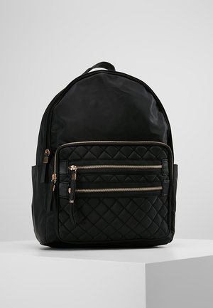 Sort rygsæk med quiltet frontlomme, to gyldne lynlåse, sidelommer og et håndtag på toppen, vist på en hvid podium mod en grå baggrund.