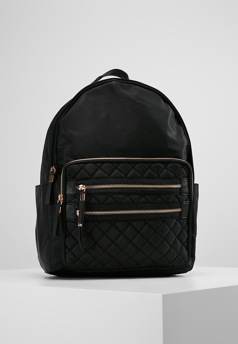 Sac à dos noir avec une poche avant matelassée, deux fermetures éclair dorées, des poches latérales et une poignée supérieure, présenté sur un piédestal blanc contre un fond gris.