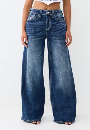 Vrouw die hoge taille, wijd uitlopende blauwe denim jeans draagt met vervaagd voorpand en witte contrasterende stiksels, staand met ontspannen handen langs haar zij.