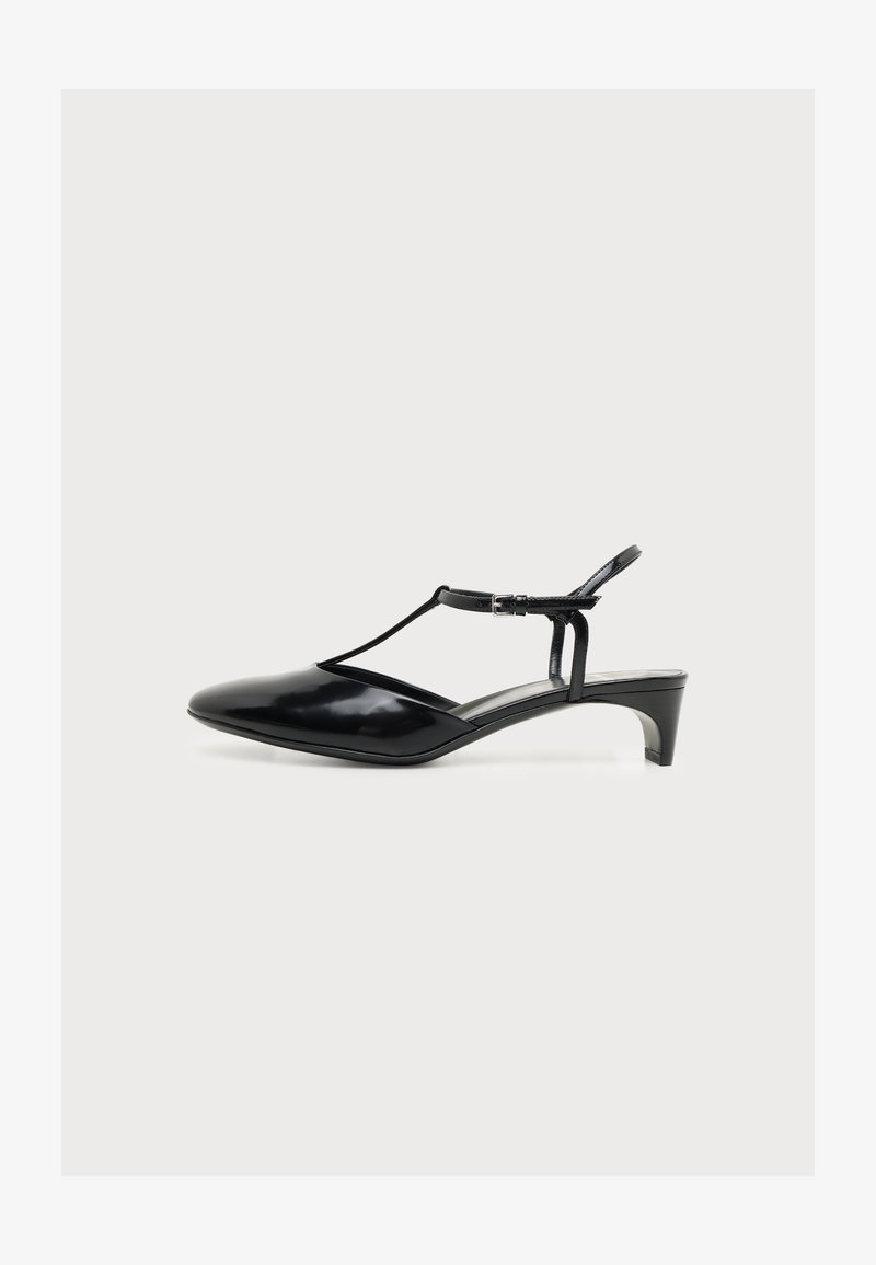 Chaussure en cuir verni noir à talon bas avec bride en T et boucle à la cheville, bout pointu et design slingback sur fond blanc.