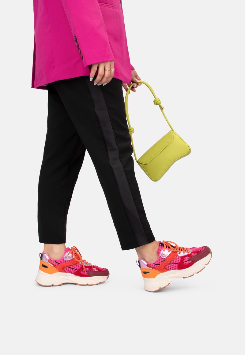 Zapatillas deportivas gruesas y multicolor con malla, detalles en rosa y naranja, combinadas con pantalones negros ajustados y un pequeño bolso verde.