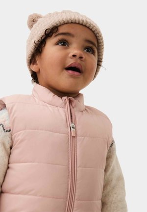 Bambino piccolo che indossa un berretto beige lavorato a maglia e un gilet trapuntato rosa chiaro, guarda verso l'alto con la bocca leggermente aperta.