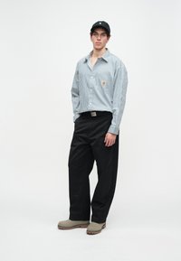 Camiseta de manga larga a rayas en azul claro con un logo en el pecho, combinada con pantalones negros holgados y zapatos tipo slip-on grises. Gorra de béisbol negra en la cabeza.